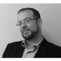 Black and white portrait of a man with glasses and beard wearing a suit jacket over a collared shirt.