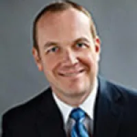 Headshot of a smiling man in a dark suit and blue tie against a gray background