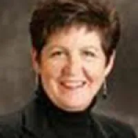 Smiling woman with short brown hair wearing a black turtleneck and earrings against a neutral background