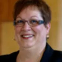 Smiling middle-aged woman with short hair and glasses wearing a black blazer and pearl earrings
